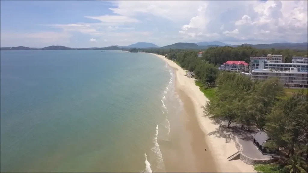 Aerial view of the long sandy beach at Laem Mae Phim, with the sea on the left, casuarina trees and low buildings along the shore and hills in the distance