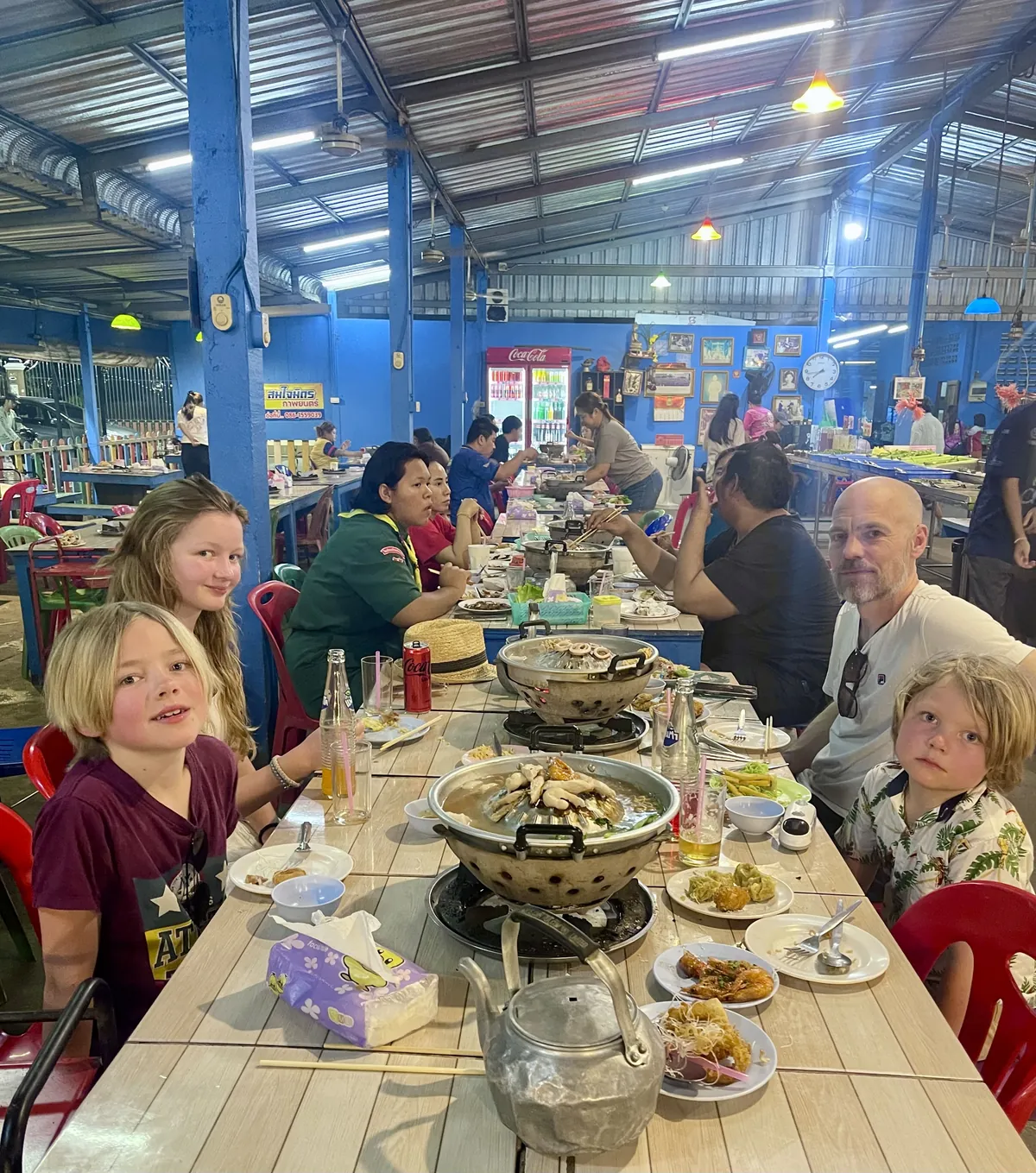 A family sharing hot-pot soup and Thai dishes at a long table in a busy open-air restaurant