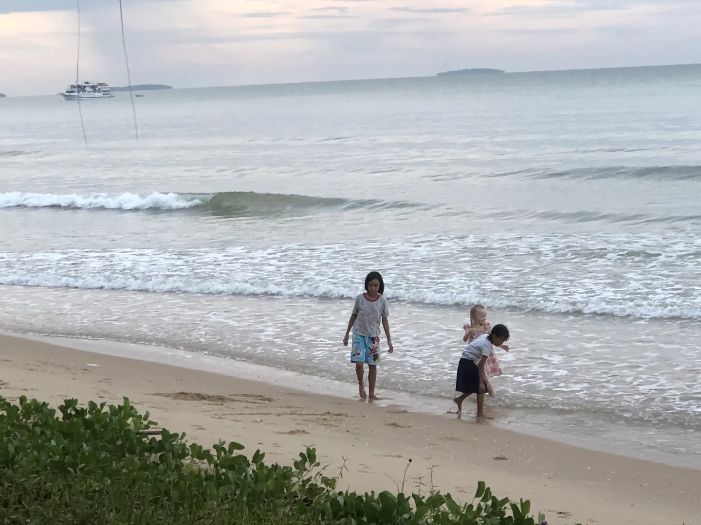 Three children paddling at the water's edge on a wide beach, with boats and islands on the horizon