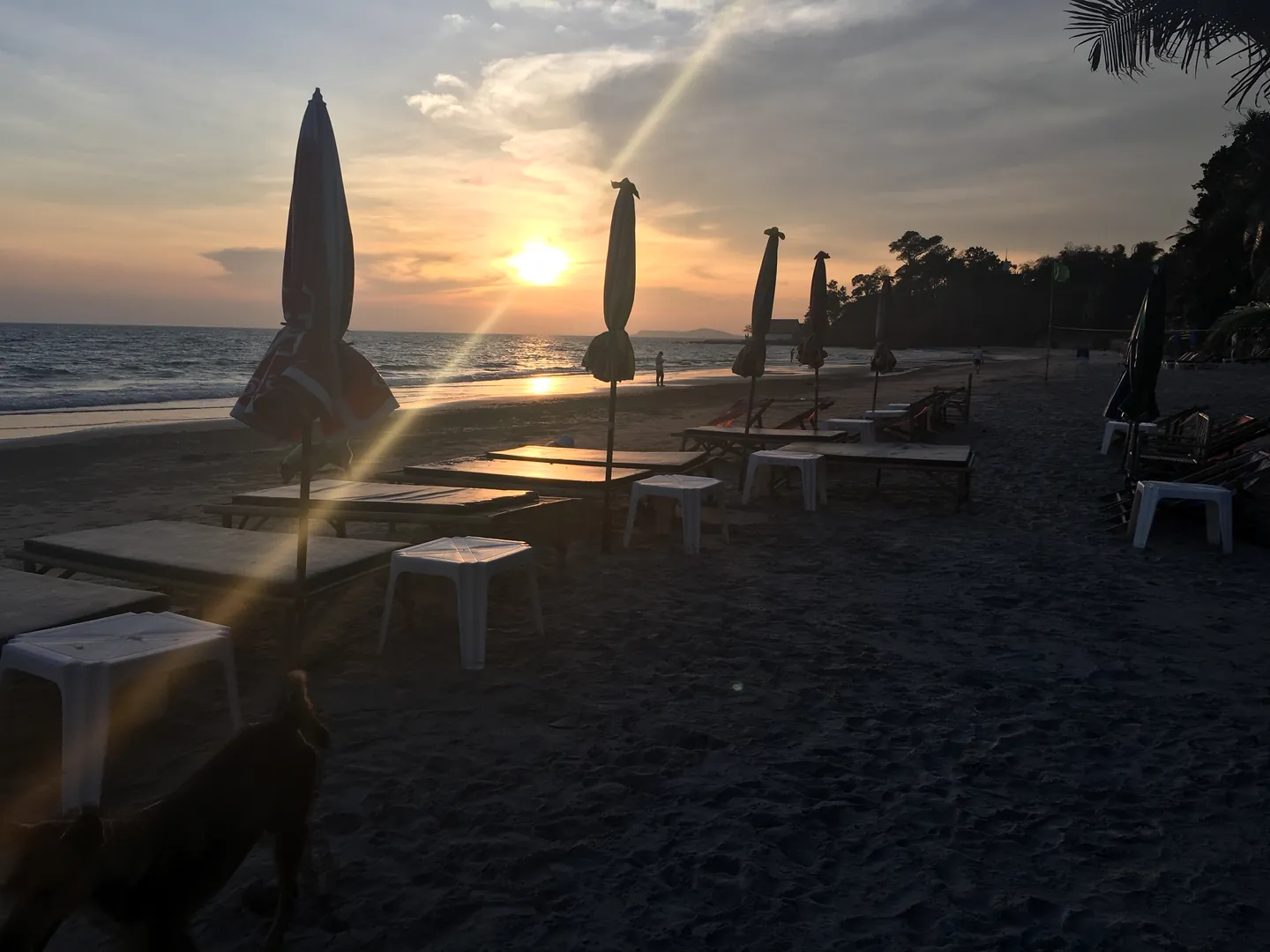 Low tables and stools set out on the sand for dinner, with closed parasols and the sun setting over the sea